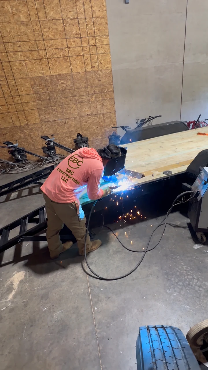 Worker in a coral EBC sweatshirt welding a black trailer frame in a workshop